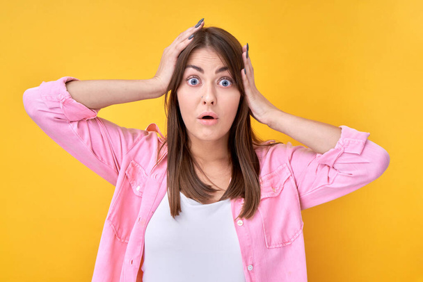 Shocked brunette girl looking surprised with open mouth holding head on yellow background with copy space - Фото, зображення