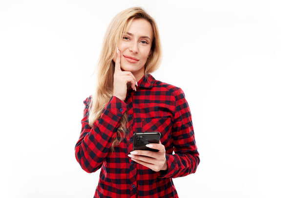 Pensive thinking blond girl in casual holding smartphone isolated on white studio background - Φωτογραφία, εικόνα