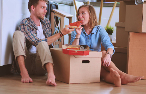 Pareja joven tiene una pausa para comer pizza en el suelo después de mudarse a una nueva casa con cajas a su alrededor. Pareja joven - Foto, Imagen