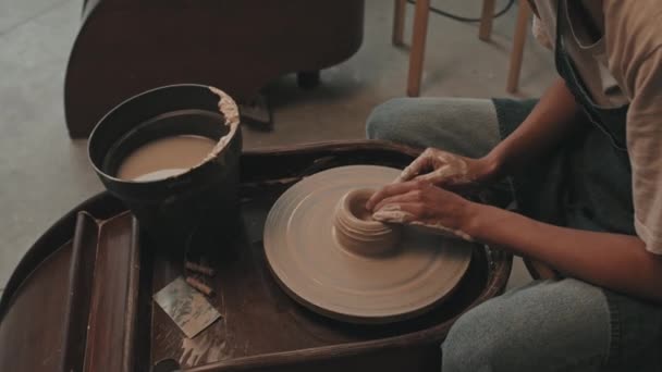 High angle side view of cropped female artist wearing apron and t-shirt, sitting in modern workshop, shaping clay on spinning pottery wheel, making ceramics - Footage, Video