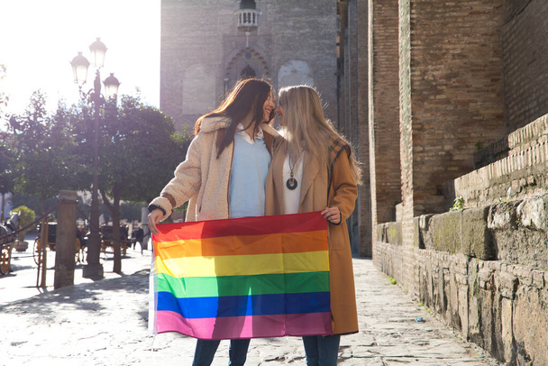 Брак лесбиянок на отдыхе и туризме в Севилье. Они стоят перед собором и во время поцелуя держат в руках флаг гей-гордости. Концепция равенства и прав lgtb. - Фото, изображение