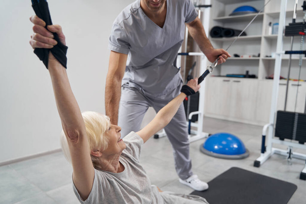 Female pensioner doing cable gym equipment exercise for rehabilitation - Foto, Imagen