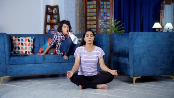 Een jonge Indiase huisvrouw die thuis yoga beoefent tijdens de lockdown periode - Pranayam poseren. Een hyperactief kind stoort zijn moeder tijdens het sporten - ongehoorzaam, luidruchtig, speels, ondeugend jongen - Video