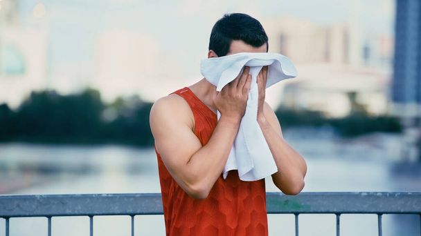 young sportsman wiping face with towel while standing on bridge - Photo, Image