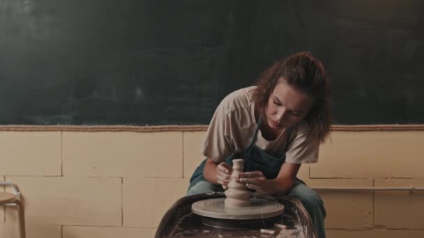 Medium lang van brunette petite blanke vrouw zitten in moderne werkplaats, het vormgeven van bizarre vaas van zachte klei op spinnende aardewerk wiel - Video