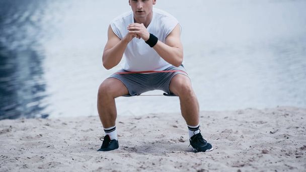cropped view of athletic sportsman doing squat exercise on river shore  - Фото, изображение