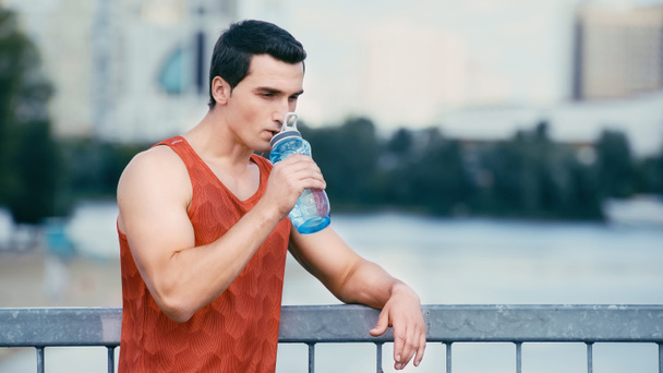 Müder Sportler steht auf Brücke und trinkt Wasser aus Sportflasche - Foto, Bild