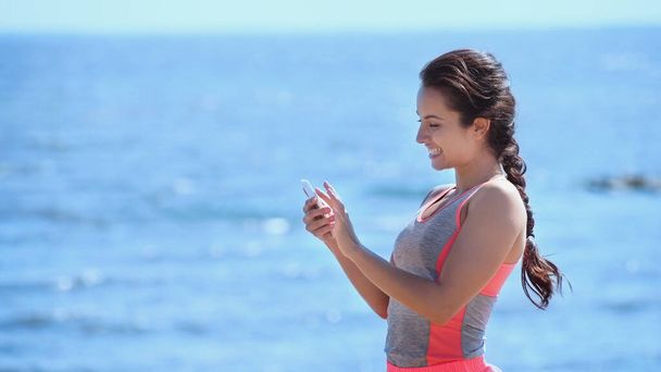 Smiling sportswoman using smartphone near blurred sea  - Photo, Image