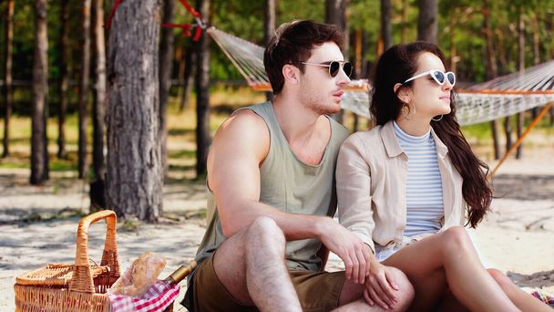 Młoda para w okularach przeciwsłonecznych siedzi przy koszyku piknikowym na plaży  - Zdjęcie, obraz
