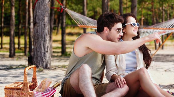 Muž v slunečních brýlích ukazuje prstem blízko usmívající se přítelkyně a piknikový koš na pláži  - Fotografie, Obrázek