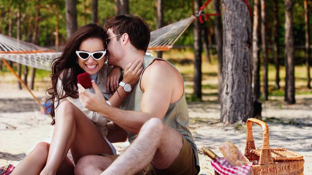 Man in sunglasses holding jewelry box near cheerful girlfriend and basket on beach  - Photo, Image