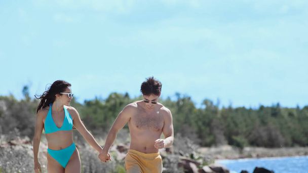 Giovane coppia in occhiali da sole e costumi da bagno a piedi sulla spiaggia  - Foto, immagini