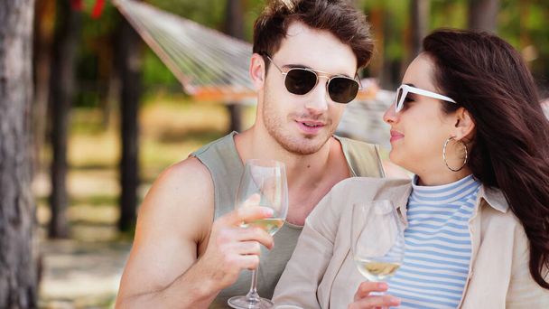 Young man in sunglasses holding glass of wine near girlfriend on beach  - Foto, afbeelding