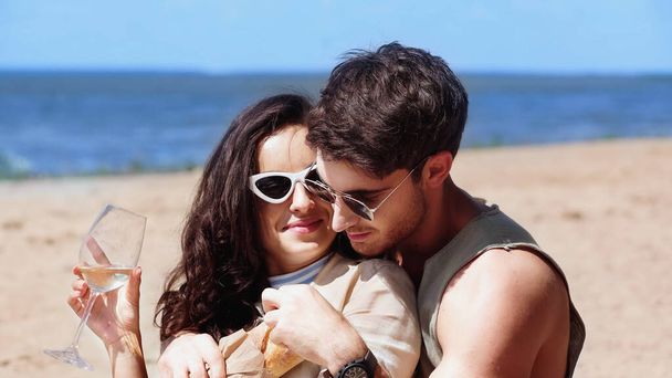 Young man hugging girlfriend in sunglasses with wine and baguette on beach  - Photo, Image