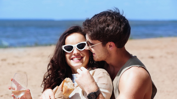 glimlachende man in zonnebril knuffelen vriendin met wijn en stokbrood op het strand  - Foto, afbeelding