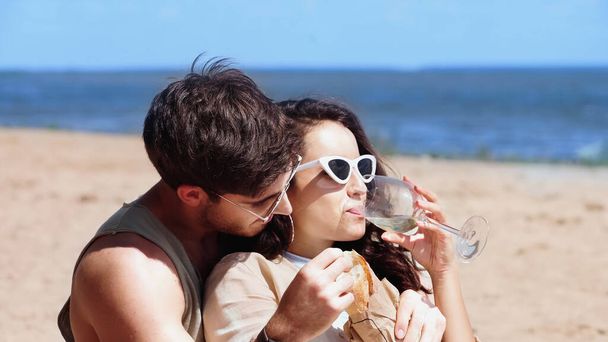 Woman in sunglasses drinking wine near boyfriend with baguette on beach  - Photo, Image