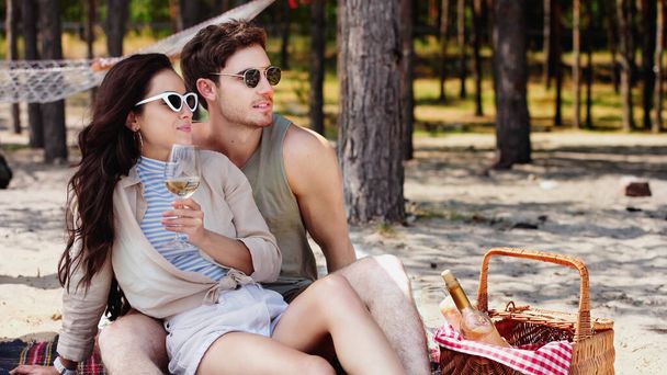Giovane donna in occhiali da sole in possesso di vino vicino fidanzato e cesto sulla spiaggia  - Foto, immagini