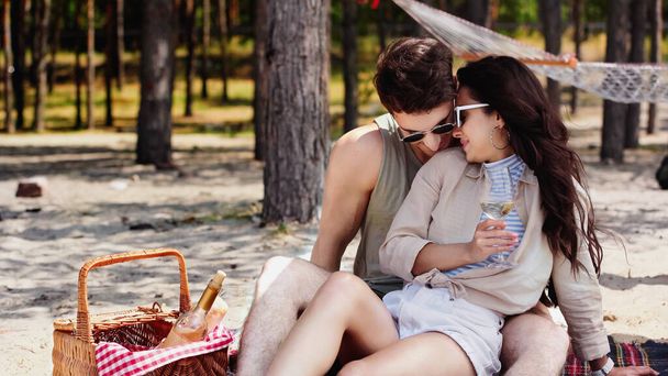 Молодая женщина в солнечных очках держит вино и сидит рядом с парнем на одеяле на пляже  - Фото, изображение