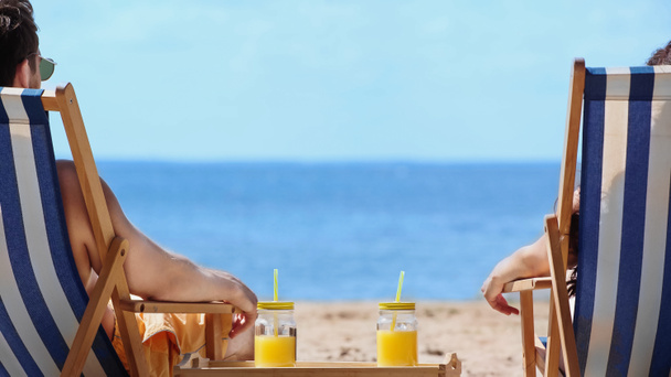 Vista trasera de pareja sentada en tumbonas cerca de zumo de naranja en la playa  - Foto, imagen