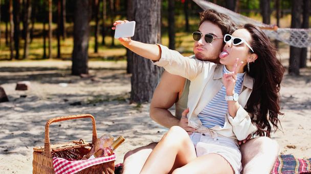 Pareja joven tomando selfie en el teléfono inteligente cerca de la cesta de picnic en la playa  - Foto, Imagen