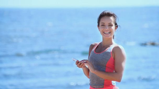 Positive woman in sportswear holding cellphone and looking at camera near blurred sea  - Фото, зображення