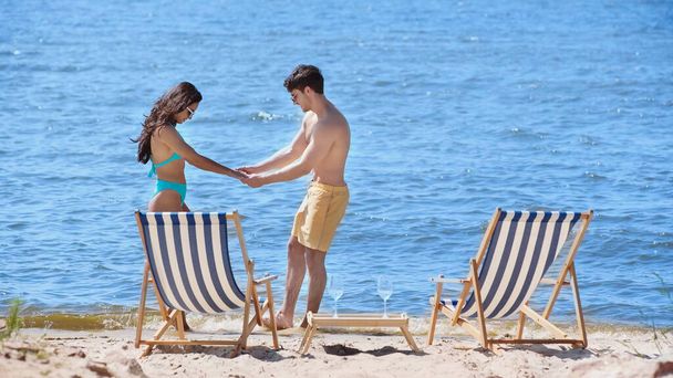 Вид збоку усміхненої пари в купальнику тримає руки біля стільців палуби та вина на пляжі
  - Фото, зображення