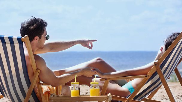 Hombre en gafas de sol cogido de la mano de la novia y señalando con el dedo cerca del jugo de naranja en la playa  - Foto, imagen