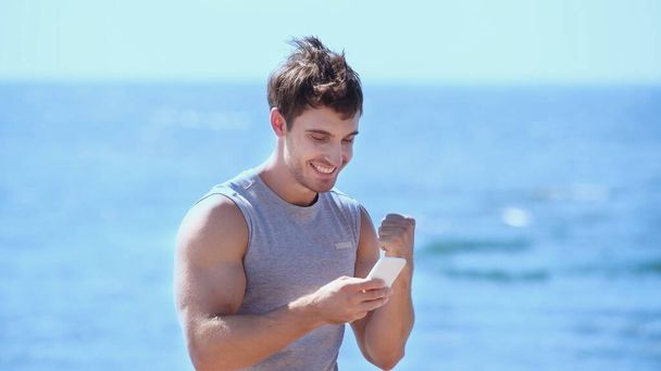 Excited sportsman using mobile phone near blurred sea  - Фото, изображение