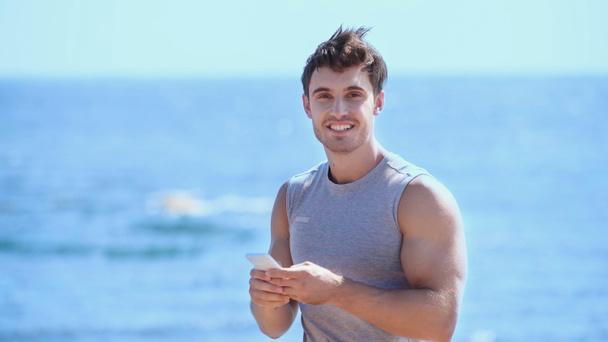 Deportista positivo usando teléfono inteligente y mirando a la cámara cerca del mar borroso  - Foto, imagen
