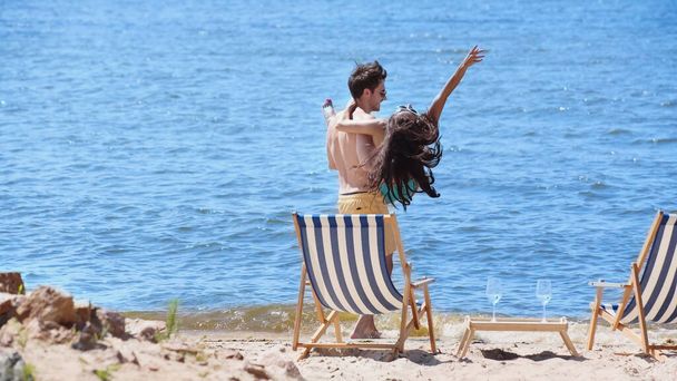 Homme positif levant petite amie près des chaises longues et du vin sur la plage  - Photo, image