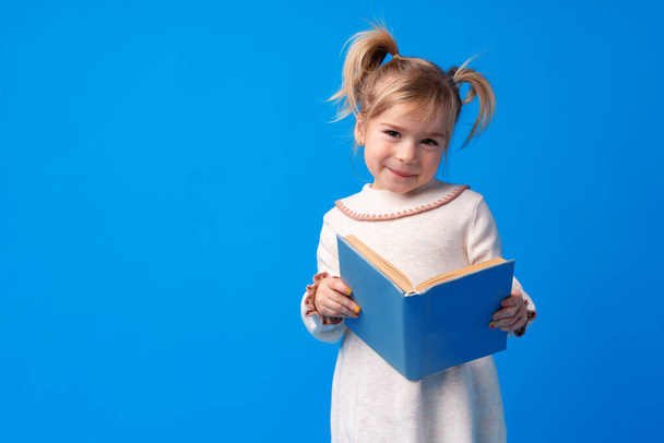 Little beautiful smiling girl holding book against blue background - Foto, afbeelding