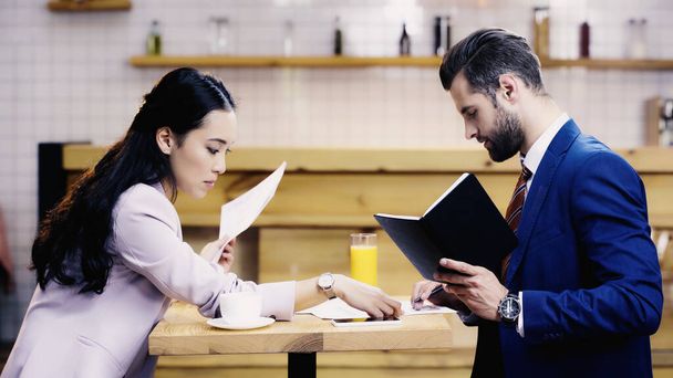 азіатська бізнес-леді дивиться на документи біля бородатого бізнесмена в кафе
 - Фото, зображення
