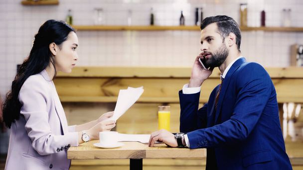 asian businesswoman holding documents near businessman talking on smartphone in cafe - Zdjęcie, obraz