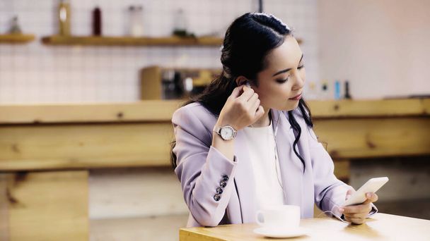 jeune femme asiatique en costume écouter de la musique dans les écouteurs et en utilisant smartphone dans le café  - Photo, image