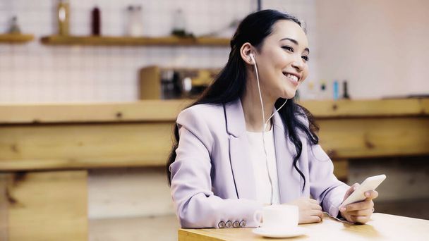 happy asian woman in suit listening music in earphones and holding mobile phone near cup in cafe - 写真・画像