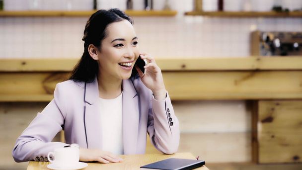 cheerful asian woman in suit talking on smartphone in cafe  - 写真・画像