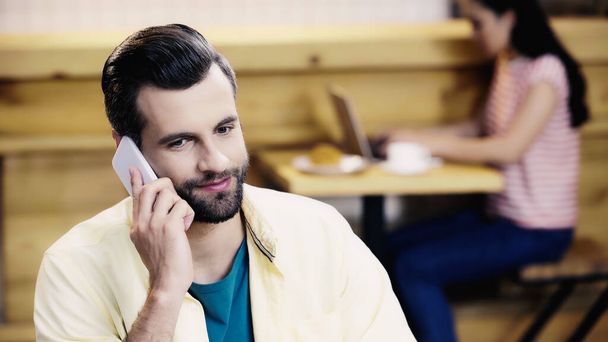 hombre barbudo hablando en el teléfono inteligente cerca de mujer borrosa en la cafetería - Foto, Imagen