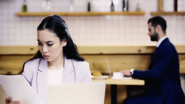 молодая азиатская предпринимательница просматривает документы рядом с размытым бизнесменом в кафе - Фото, изображение