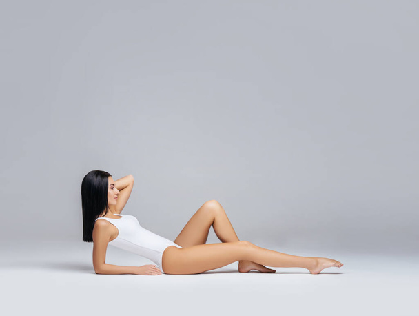 Young, fit and beautiful brunette woman in white swimsuit posing in studio. Concept of fitness, dieting and skin care. - Foto, Imagen