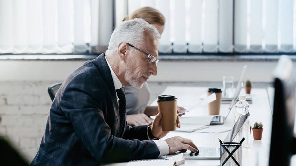 Вид сбоку зрелого бизнесмена, держащего кофе и использующего ноутбук в офисе  - Фото, изображение