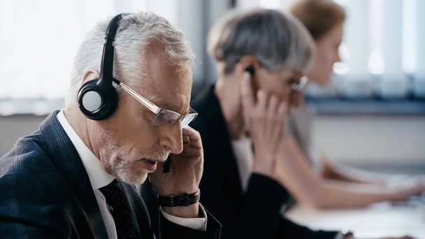 Ώριμος επιχειρηματίας με ακουστικά που μιλάει κοντά σε θολή συναδέλφους στο γραφείο  - Φωτογραφία, εικόνα