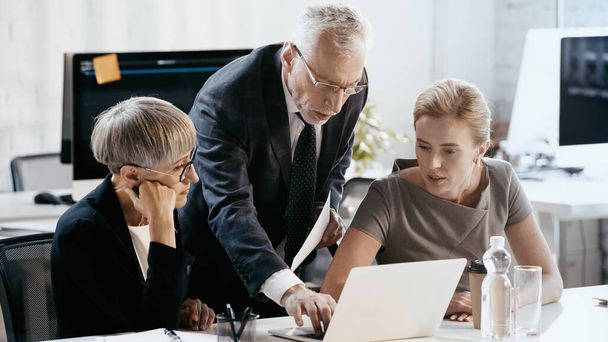 Зрелый бизнесмен, использующий ноутбук рядом с коллегами в офисе  - Фото, изображение