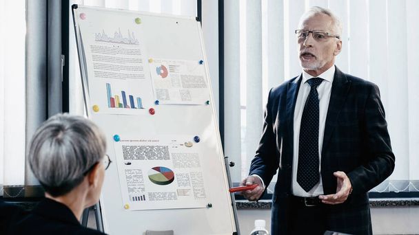 Mature businessman in formal wear talking near flip chart and colleague in office  - Photo, Image