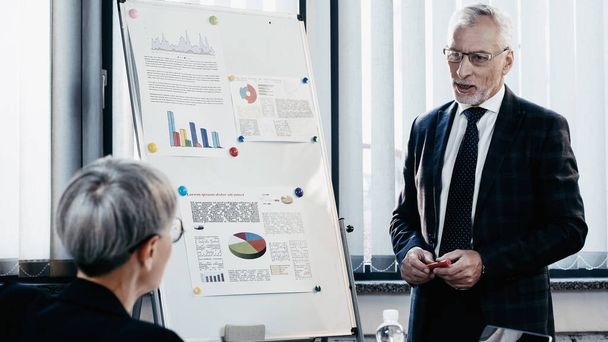 Middle aged businessman talking near blurred businesswoman and flip chart in office  - Photo, Image