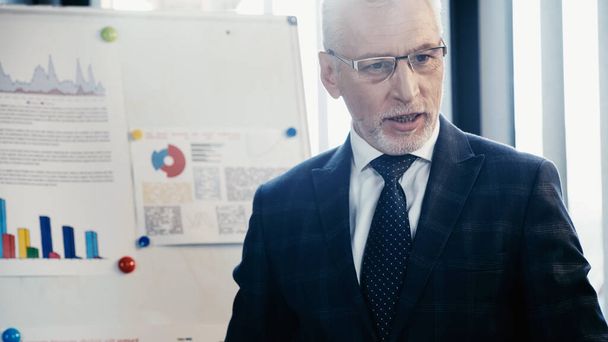 Uomo d'affari maturo in abbigliamento formale e occhiali in piedi vicino a flip chart in ufficio  - Foto, immagini