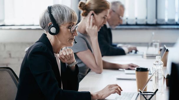 Side view of mature businesswoman in headset using laptop near blurred colleagues in office  - Фото, изображение
