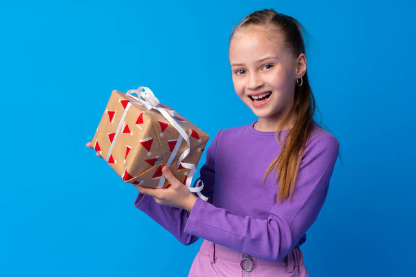 Happy little girl with long hair holding a gift box on a blue background - 写真・画像
