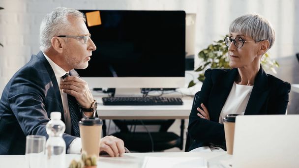 Зрелый бизнесмен разговаривает с коллегой возле бумажных стаканчиков и ноутбука в офисе  - Фото, изображение