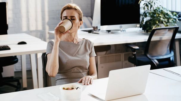 Businesswoman drinking coffee to go near takeaway salad and laptop in office  - Fotó, kép