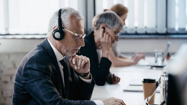 Middle aged businessman in headset using laptop near drinks in office  - Foto, imagen